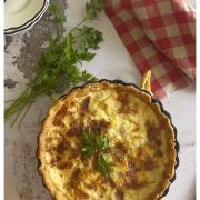 A baked quiche sits on a light surface, garnished with parsley. Nearby are a red-and-white checkered cloth, fresh parsley, a sliced orange, and stacked plates. Text reads: Quiche Lorraine, Easy Classic Recipe.