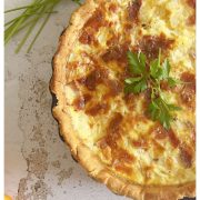 A baked quiche Lorraine in a tart pan garnished with parsley, set on a rustic white surface with fresh herbs and an orange slice nearby. Text reads: Simple French Quiche Lorraine.