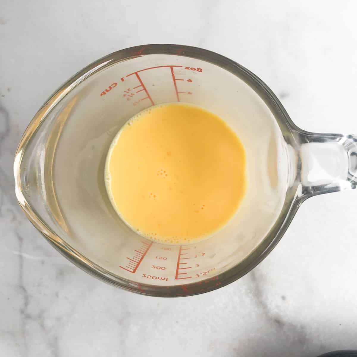 A glass measuring cup containing a small amount of beaten egg mixture, perfect for preparing a Mediterranean quiche, sits on a white marble surface.