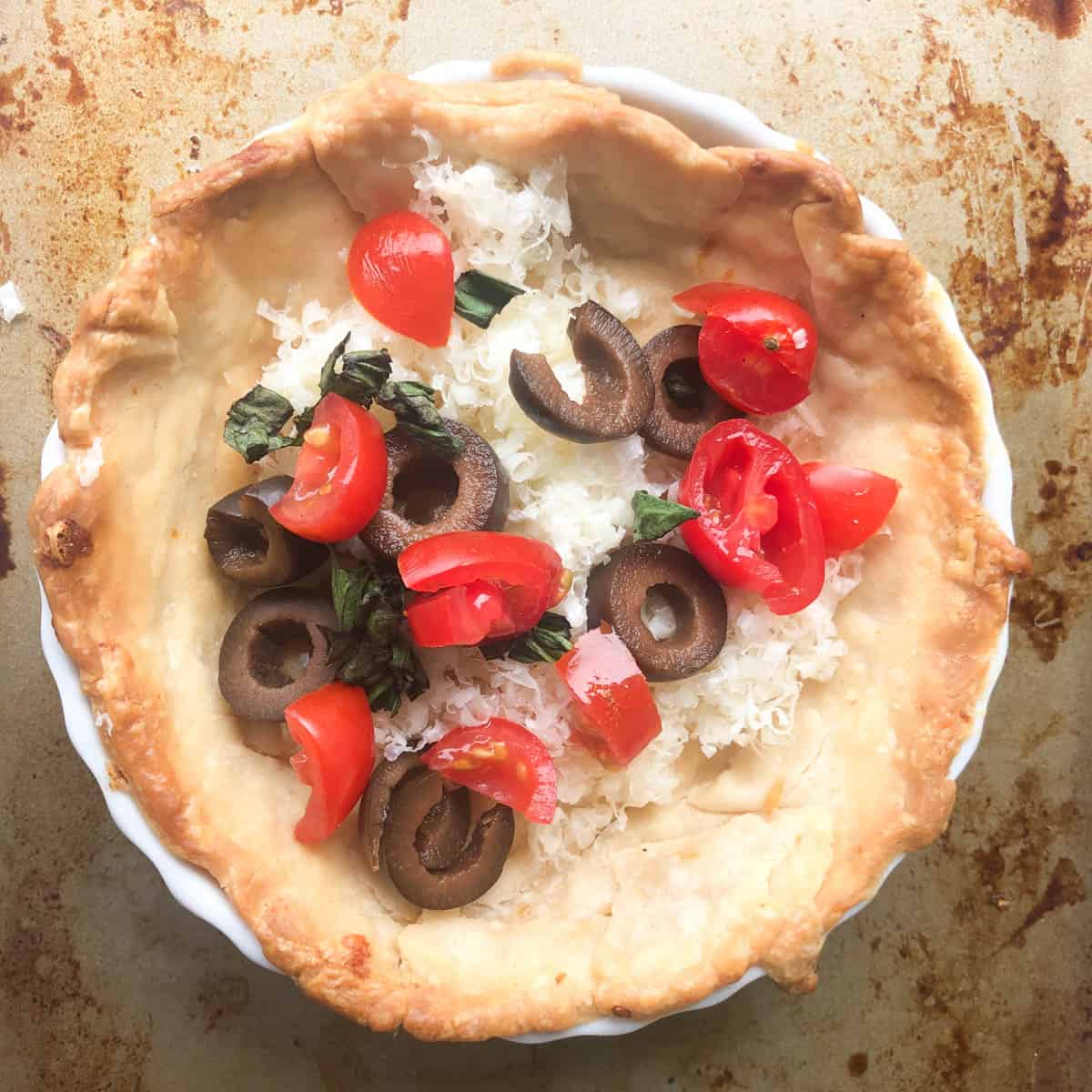 A partially baked pie crust in a white dish filled with shredded cheese, sliced black olives, chopped fresh basil, and diced cherry tomatoes—perfect for creating a vibrant Mediterranean quiche on a rustic baking tray.