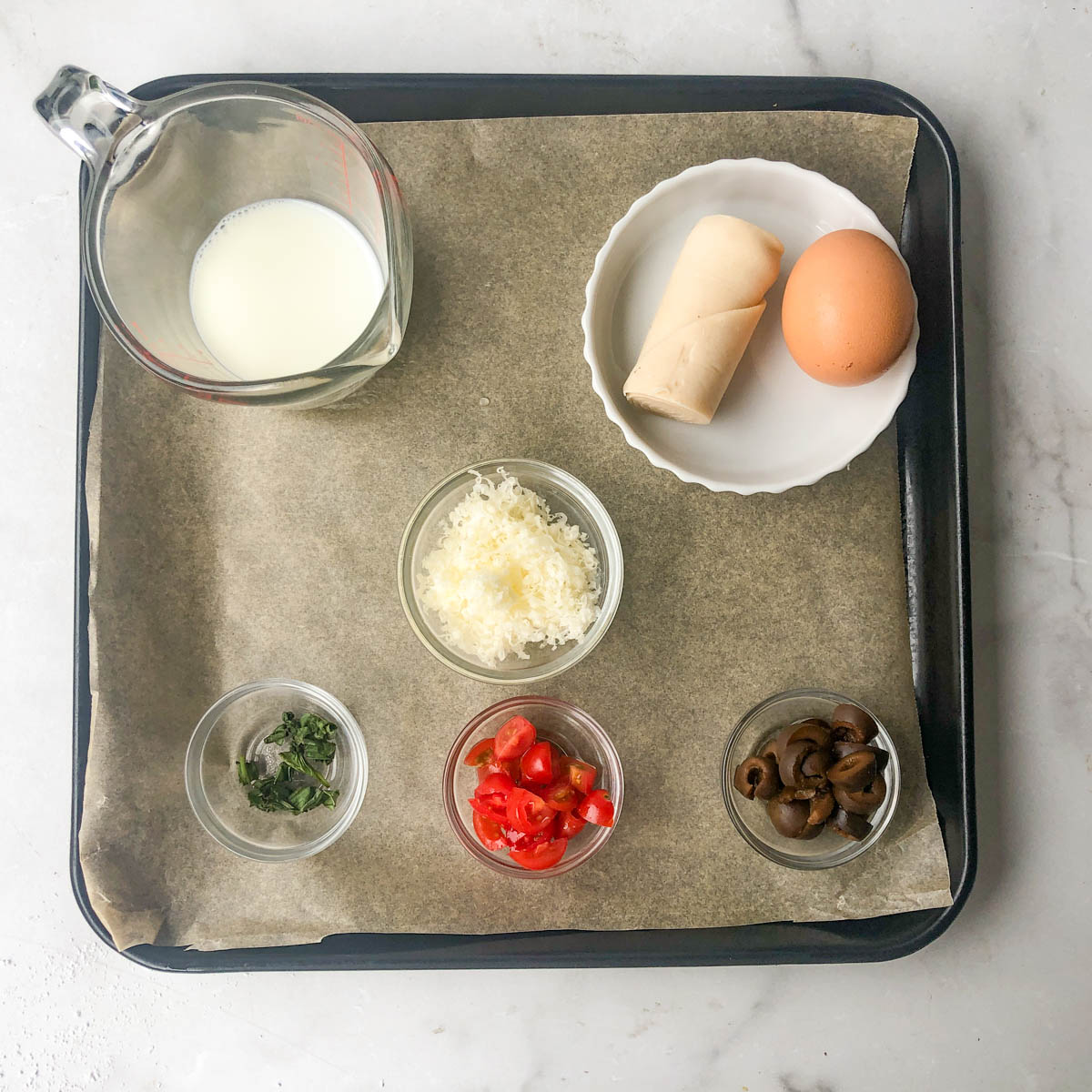 Baking tray with parchment paper holds ingredients for a mediterranean quiche: a measuring cup of milk, an egg, dough roll, shredded cheese, chopped tomatoes, sliced olives, and chopped herbs in separate bowls.