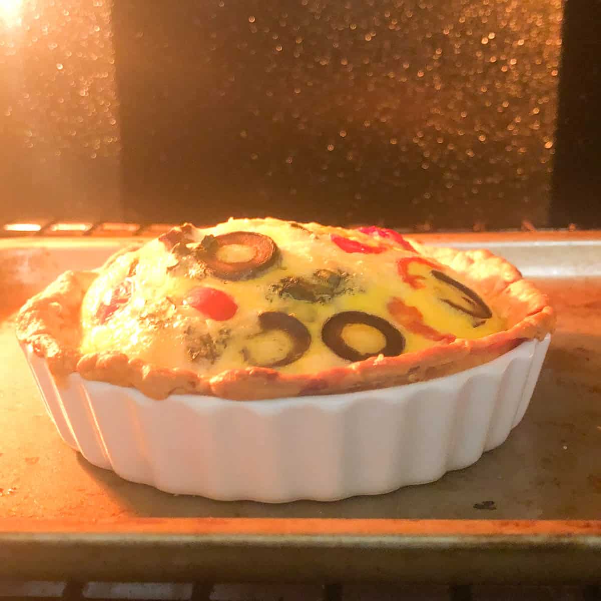A Mediterranean quiche with sliced black olives and vegetables is baking in a white ceramic dish inside an oven, its golden crust just beginning to brown.