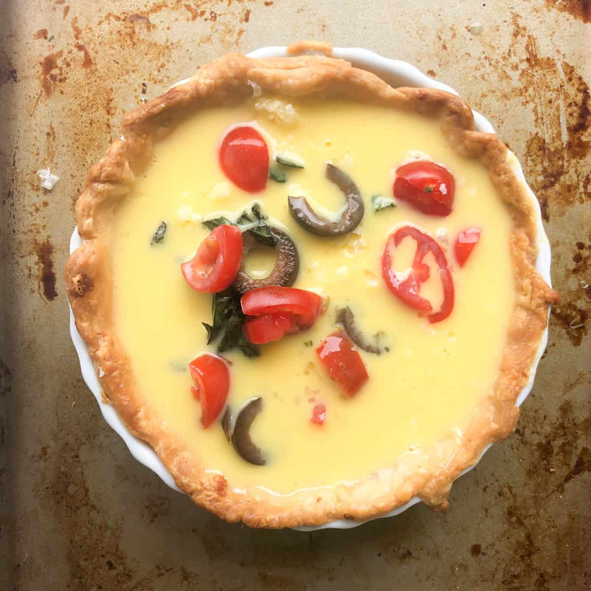A baked Mediterranean quiche with a golden crust, filled with yellow custard, halved cherry tomatoes, sliced black olives, and fresh herbs, displayed on a metal baking sheet.