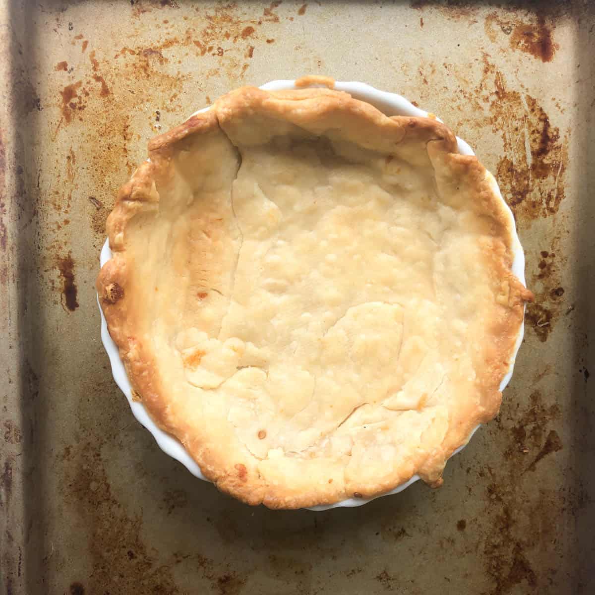 A baked pie crust for a Mediterranean quiche sits in a white dish on a worn, stained baking sheet. The golden brown crust has uneven, crimped edges and a few cracks on the surface.