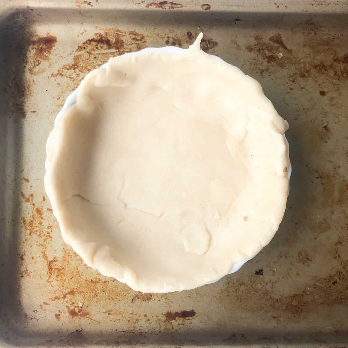 A round pie dish covered with an uncooked crust, ready for a savory Mediterranean quiche, sits on a stained metal baking sheet. The doughs edges are uneven and gently overhang the dish.