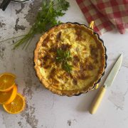A baked quiche garnished with fresh parsley sits on a marble surface beside a bunch of parsley, a knife with a cream handle, orange slices, and a red checkered cloth.