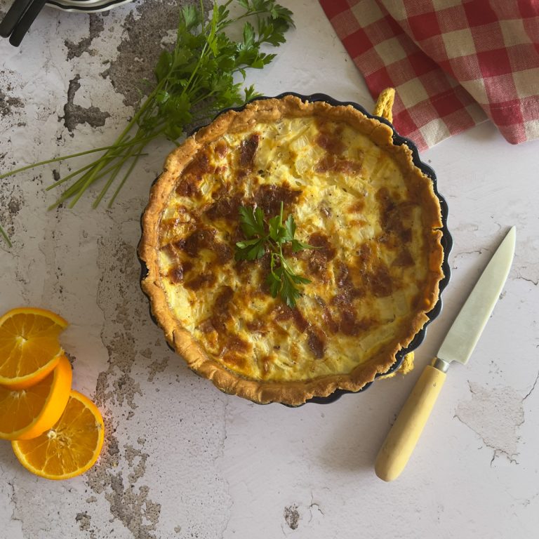 A baked quiche garnished with fresh parsley sits on a marble surface beside a bunch of parsley, a knife with a cream handle, orange slices, and a red checkered cloth.
