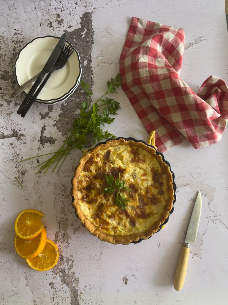 A baked quiche garnished with herbs sits on a textured surface beside a knife, three orange slices, fresh parsley, stacked plates with cutlery, and a red-and-white checkered cloth.