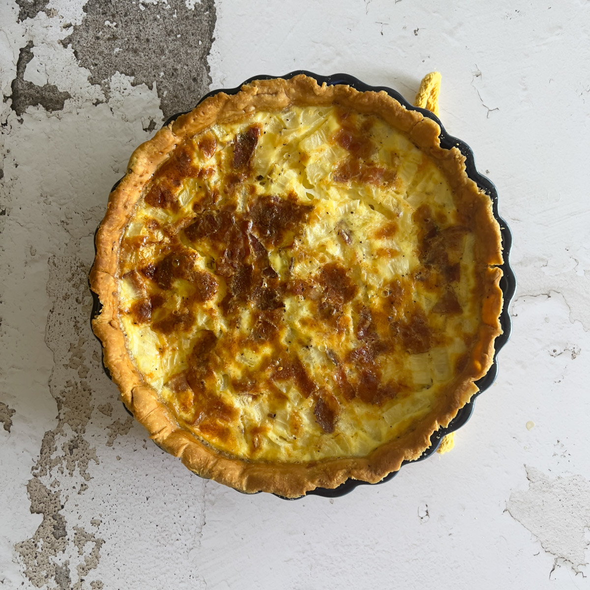 A freshly baked quiche with a golden-brown crust sits in a round black baking dish on a white, slightly cracked textured surface.