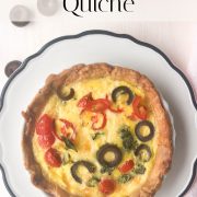 A baked individual mediterranean quiche with tomato and olive on a white plate with a decorative border, photographed from above. Text above the quiche reads INDIVIDUAL Tomato & Olive Quiche by howtomakequiche.com.
