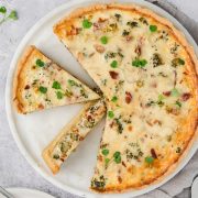 A baked quiche with broccoli, cheese, and diced ham on a white plate, garnished with microgreens. One slice is removed and placed nearby, showing the creamy filling and golden crust. A fork and napkin sit by the plate.