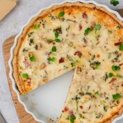 A baked quiche with a golden crust sits on a wooden board, topped with green microgreens. One large slice has been removed, showing a creamy, vegetable and ham filling. A knife and some cheese are nearby.