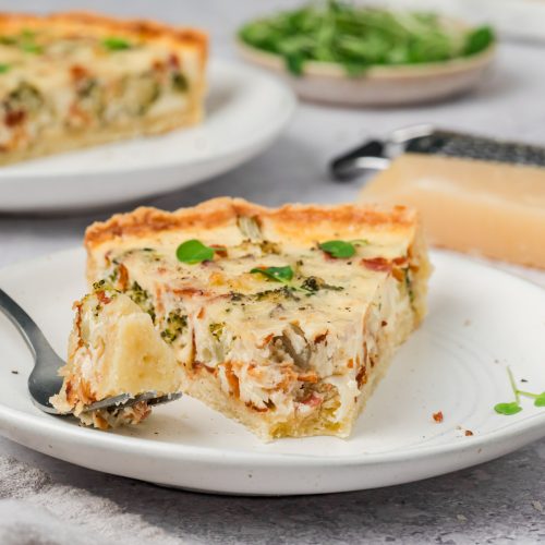A slice of savory quiche with herbs and vegetables sits on a white plate with a fork. A wedge of cheese and additional quiche are visible in the background.