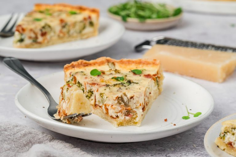 A slice of savory quiche with herbs and vegetables sits on a white plate with a fork. A wedge of cheese and additional quiche are visible in the background.
