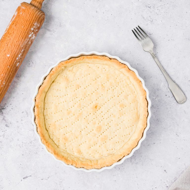 A baked, empty quiche crust in a fluted white dish sits on a light surface next to a metal fork and a wooden rolling pin dusted with flour.