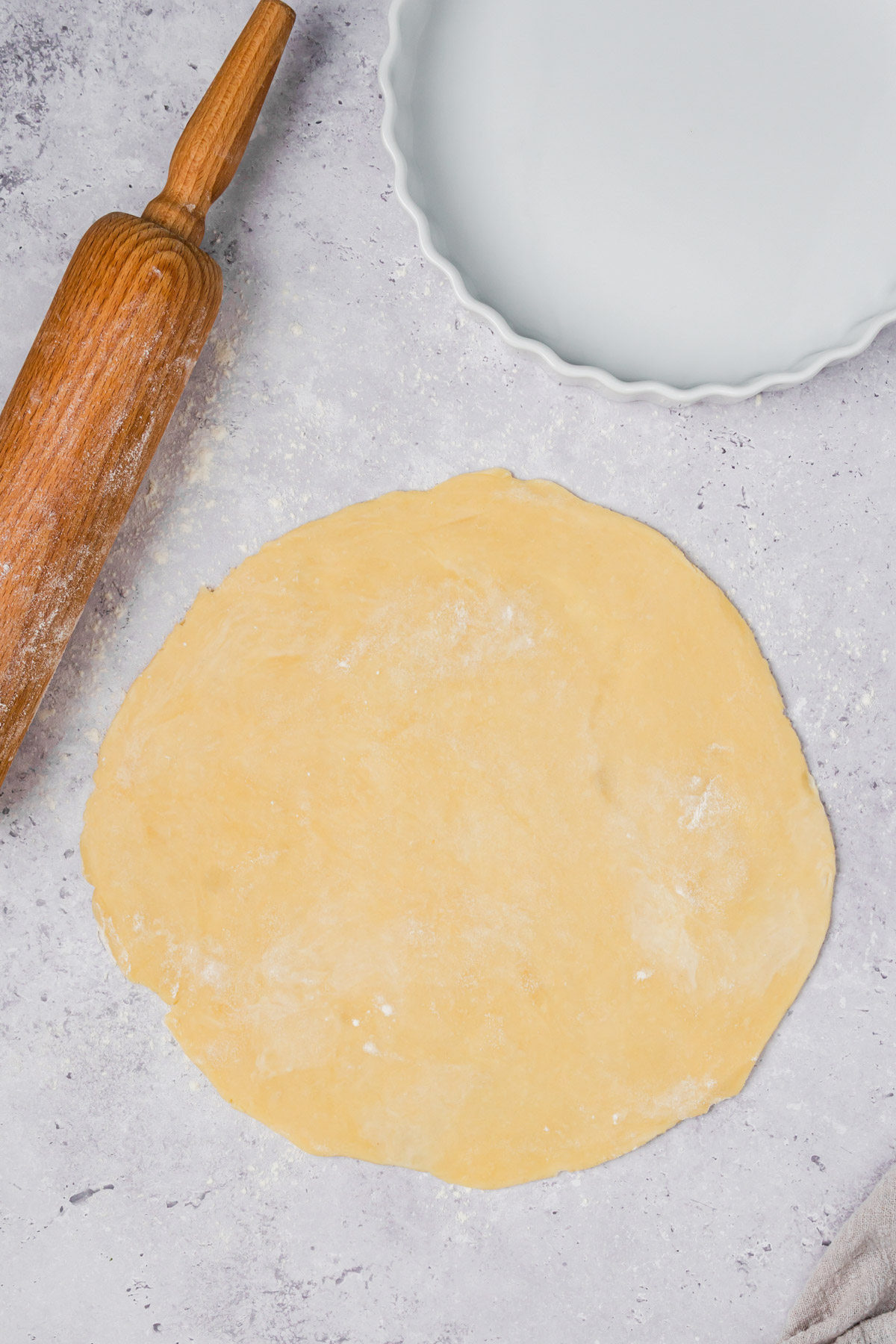 A round sheet of unbaked quiche crust dough sits on a floured surface beside a wooden rolling pin and an empty fluted-edge pie dish.