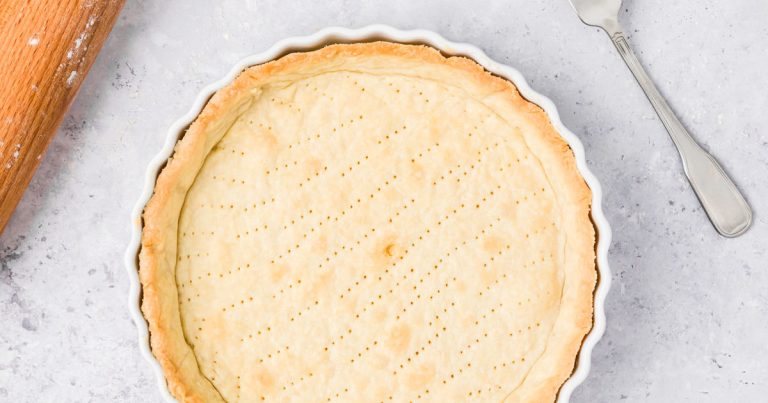 A basic quiche crust, golden and evenly pierced with small fork holes, rests in a white fluted dish on a light surface beside a fork and a flour-dusted wooden rolling pin.