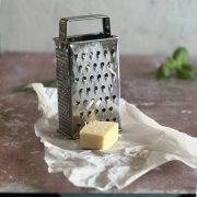 A metal box grater with some cheese remnants sits on a piece of white parchment paper next to a block of cheese, on a pinkish countertop with a green herb in the background.