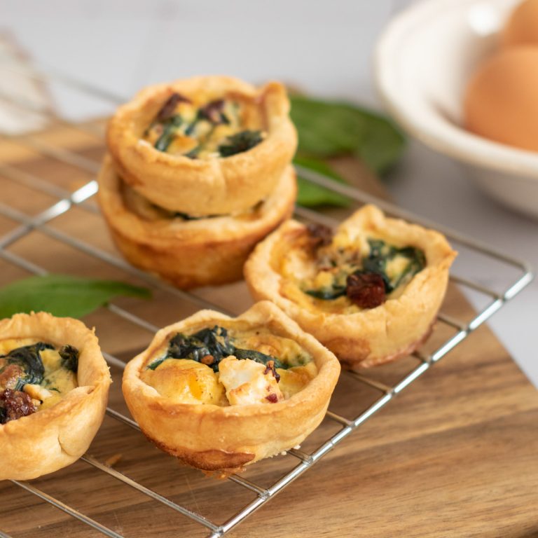 Four mini quiches with spinach and sun-dried tomatoes rest on a wire rack atop a wooden board. In the background, a bowl with brown eggs is partially visible.