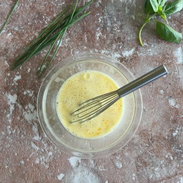A metal whisk rests in a glass bowl filled with beaten eggs on a textured brown surface. Fresh chives and basil leaves are arranged nearby for garnish.