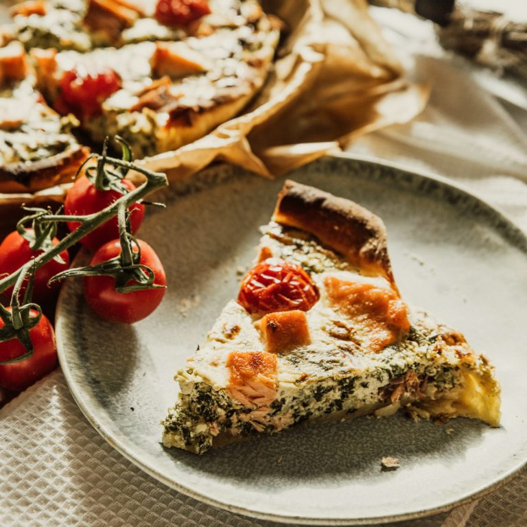 A slice of savory pie with spinach, cheese, and cherry tomatoes on a ceramic plate, with vine tomatoes beside it and more pie in the background on parchment paper.