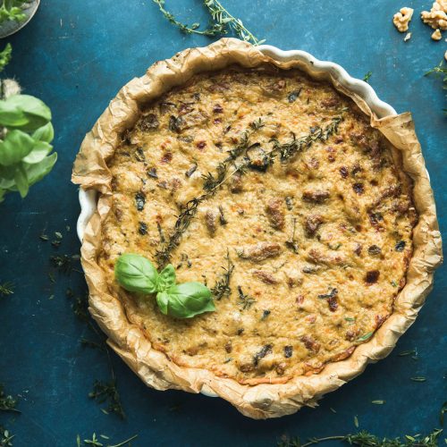 A baked savory tart in a parchment-lined dish, garnished with fresh basil leaves and thyme sprigs, sits on a blue surface with scattered herbs and walnuts nearby.