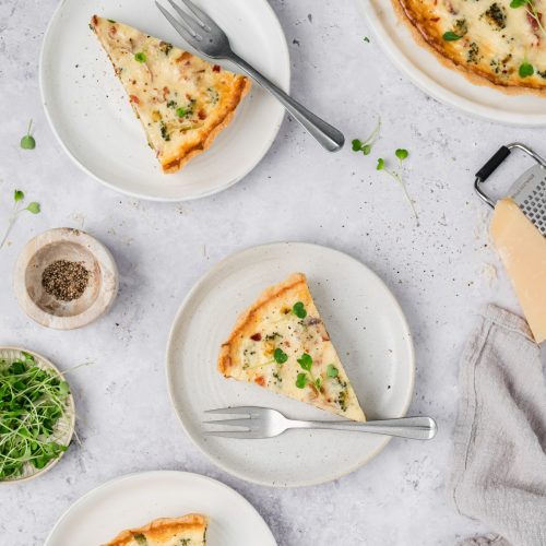 Three plates with slices of quiche, each with a fork, are arranged on a light surface with scattered microgreens, a wooden bowl of pepper, cheese, and a grater nearby.