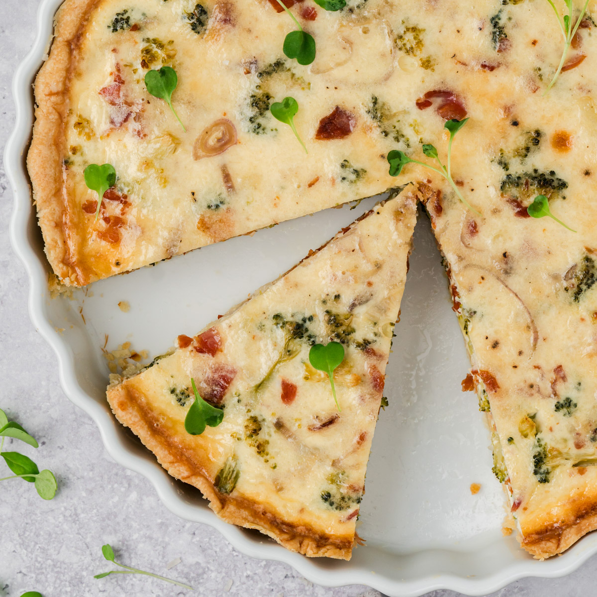 A baked quiche with a golden crust, filled with broccoli, bacon, cheese, and herbs, sits in a white pie dish. One slice is slightly removed. Small green microgreens are scattered on top.