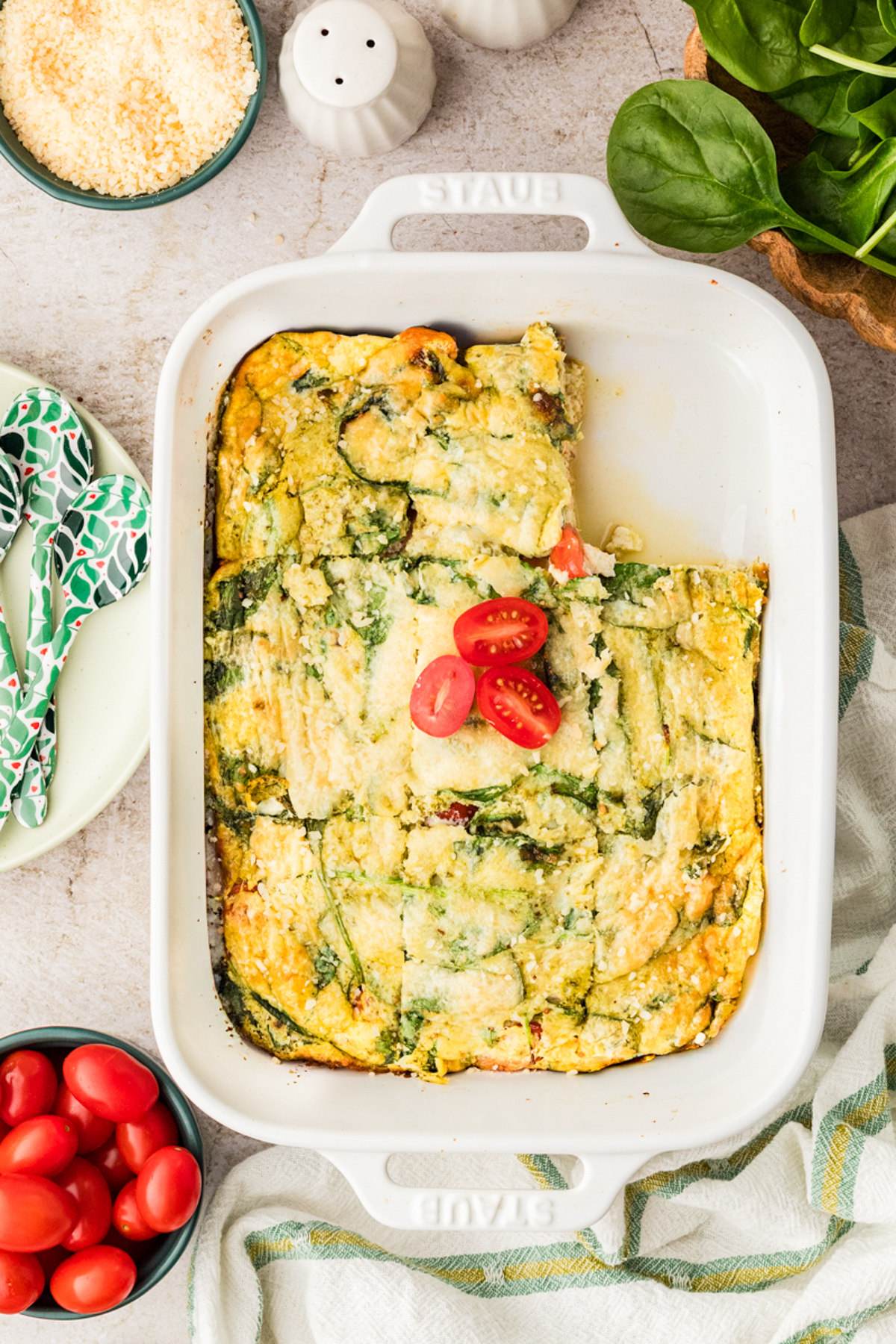 A baked egg casserole with spinach and cheese in a white dish, topped with sliced cherry tomatoes. A bowl of fresh spinach, a bowl of cherry tomatoes, and a small dish of grated cheese are nearby.