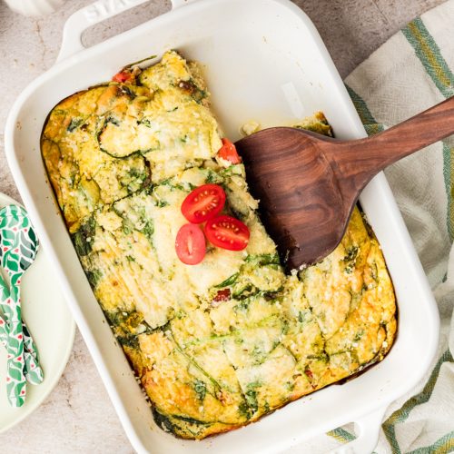 A baked egg casserole with spinach, tomatoes, and cheese in a white dish, partially sliced with a wooden spatula. Fresh spinach and a bowl of cherry tomatoes are nearby on the table.