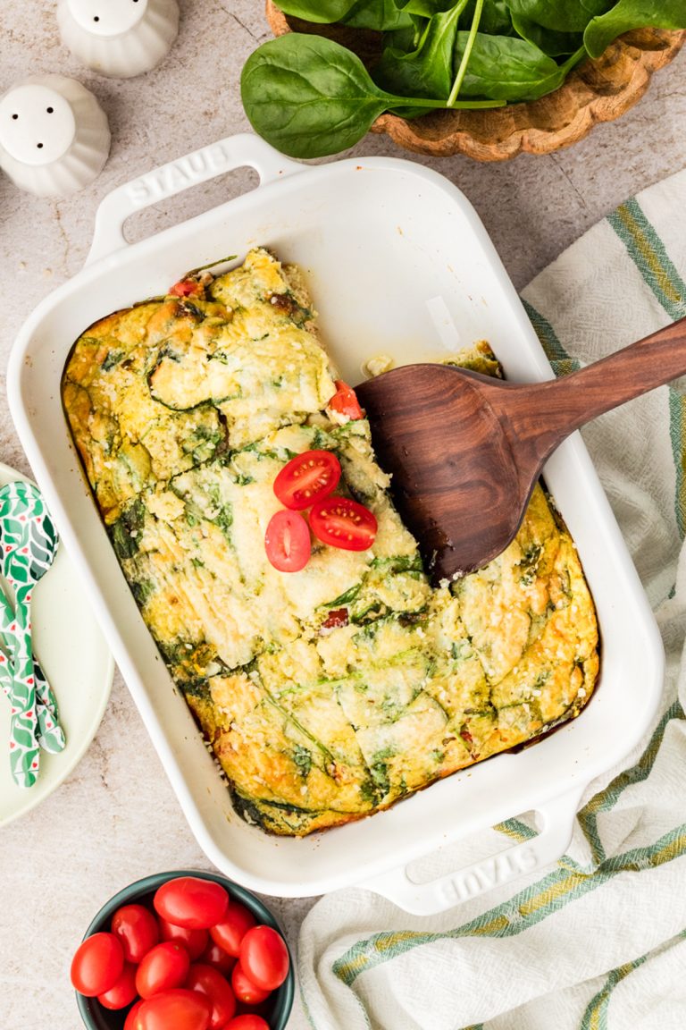 A baked egg casserole with spinach, tomatoes, and cheese in a white dish, partially sliced with a wooden spatula. Fresh spinach and a bowl of cherry tomatoes are nearby on the table.
