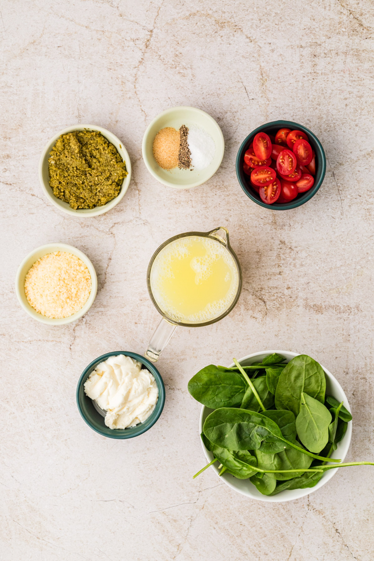 Six small bowls on a light countertop, containing pesto, grated cheese, cherry tomato halves, mayonnaise, baby spinach leaves, and a cup of broth with seasonings.