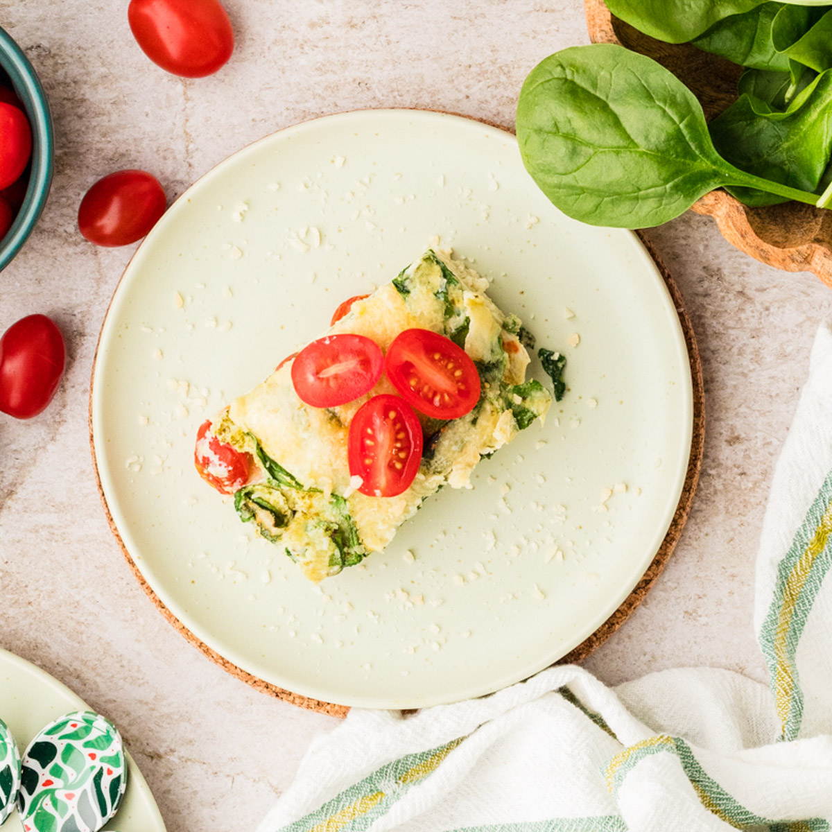 A serving of spinach and tomato lasagna roll-up topped with sliced cherry tomatoes on a light plate, surrounded by fresh spinach leaves and cherry tomatoes on a neutral surface.