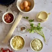 Assorted quiche ingredients on a countertop: eggs, cream, chopped bacon, fresh parsley, shredded cheese, diced onions, seasoning in a small bowl, and a roll of pie crust.
