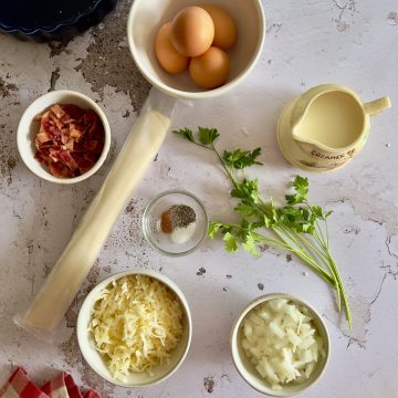 Assorted quiche ingredients on a countertop: eggs, cream, chopped bacon, fresh parsley, shredded cheese, diced onions, seasoning in a small bowl, and a roll of pie crust.